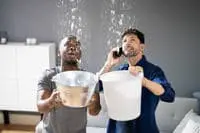 Two men are holding buckets and collecting water underneath from the leaking roof